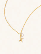 A gold chain necklace with the caplital letter X approximately 8mm x 6mm. Set on a cream background with gentle shadowing.