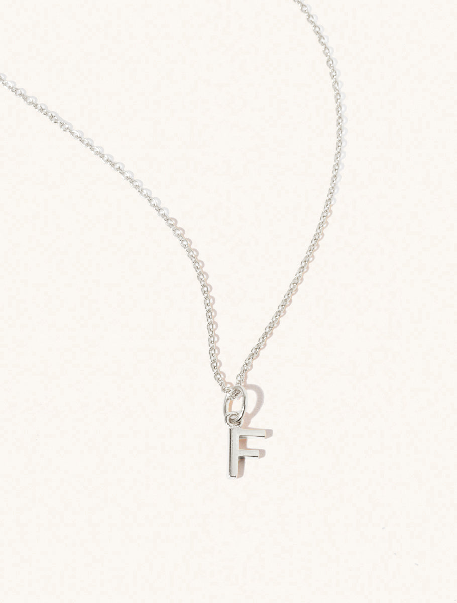 A sterling silver chain necklace with the caplital letter F approximately 8mm x 6mm. Set on a cream background with gentle shadowing.