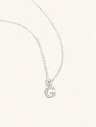 A sterling silver chain necklace with the caplital letter G approximately 8mm x 6mm. Set on a cream background with gentle shadowing.