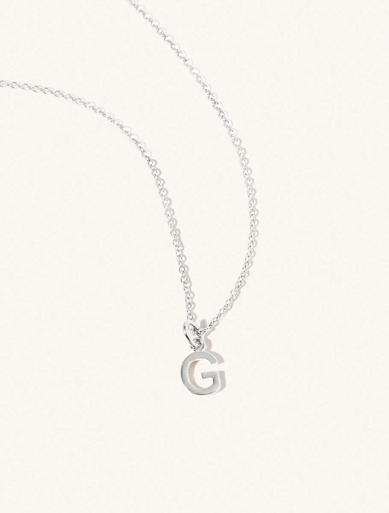 A sterling silver chain necklace with the caplital letter G approximately 8mm x 6mm. Set on a cream background with gentle shadowing.