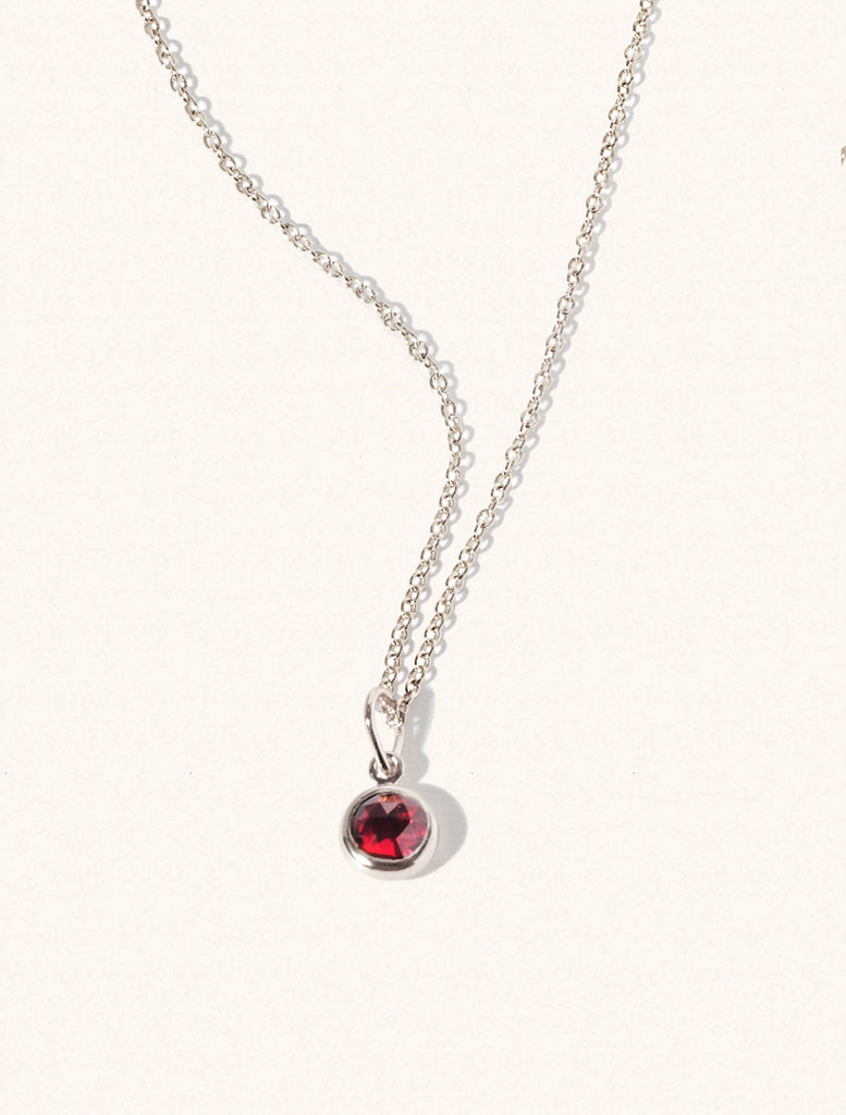 A sterling silver delicate chain nacklace with an 8mm deep red Garnet gemstone set in a round pendant. Set on an cream background with gentle shadowing.