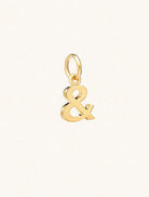 A gold & symbol approximately 8mm x 6mm with a connecting ring to thread through a necklace at the top. Set on a cream background.