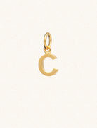 A gold caplital letter C approximately 8mm x 6mm with a connecting ring to thread through a necklace at the top. Set on a cream background.