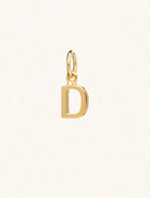 A gold caplital letter D approximately 8mm x 6mm with a connecting ring to thread through a necklace at the top. Set on a cream background.