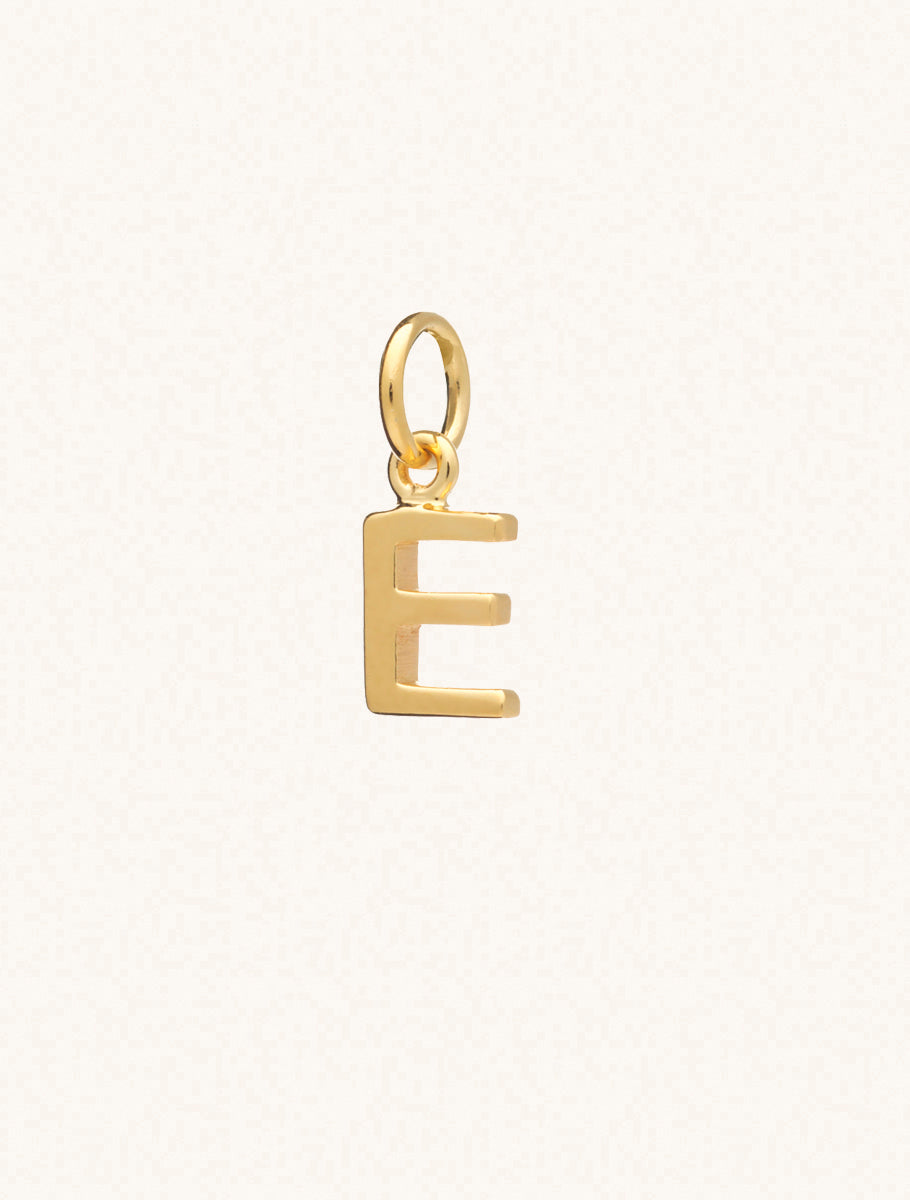 A gold caplital letter E approximately 8mm x 6mm with a connecting ring to thread through a necklace at the top. Set on a cream background.