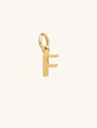 A gold caplital letter F approximately 8mm x 6mm with a connecting ring to thread through a necklace at the top. Set on a cream background.