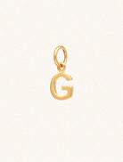 A gold caplital letter G approximately 8mm x 6mm with a connecting ring to thread through a necklace at the top. Set on a cream background.