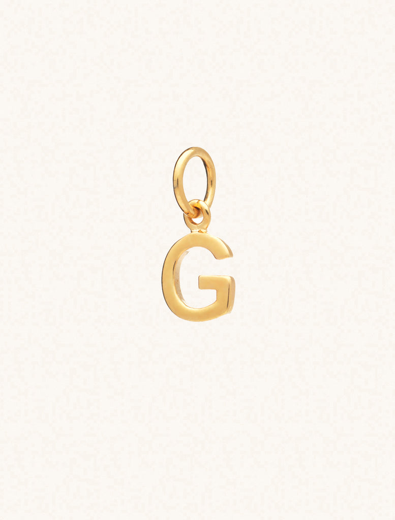 A gold caplital letter G approximately 8mm x 6mm with a connecting ring to thread through a necklace at the top. Set on a cream background.