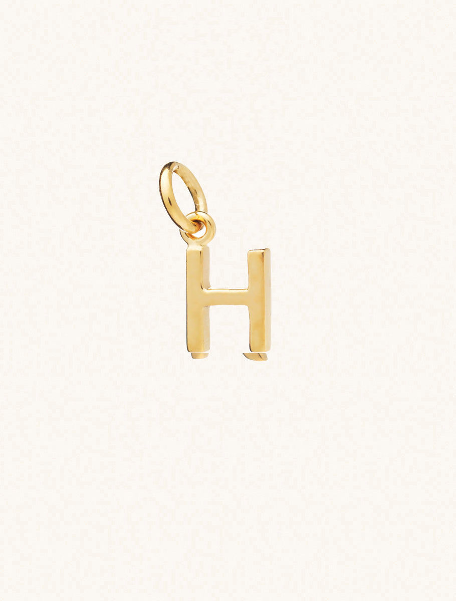 A gold caplital letter H approximately 8mm x 6mm with a connecting ring to thread through a necklace at the top. Set on a cream background.