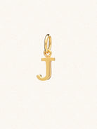 A gold caplital letter J approximately 8mm x 6mm with a connecting ring to thread through a necklace at the top. Set on a cream background.