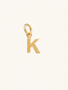 A gold caplital letter K approximately 8mm x 6mm with a connecting ring to thread through a necklace at the top. Set on a cream background.