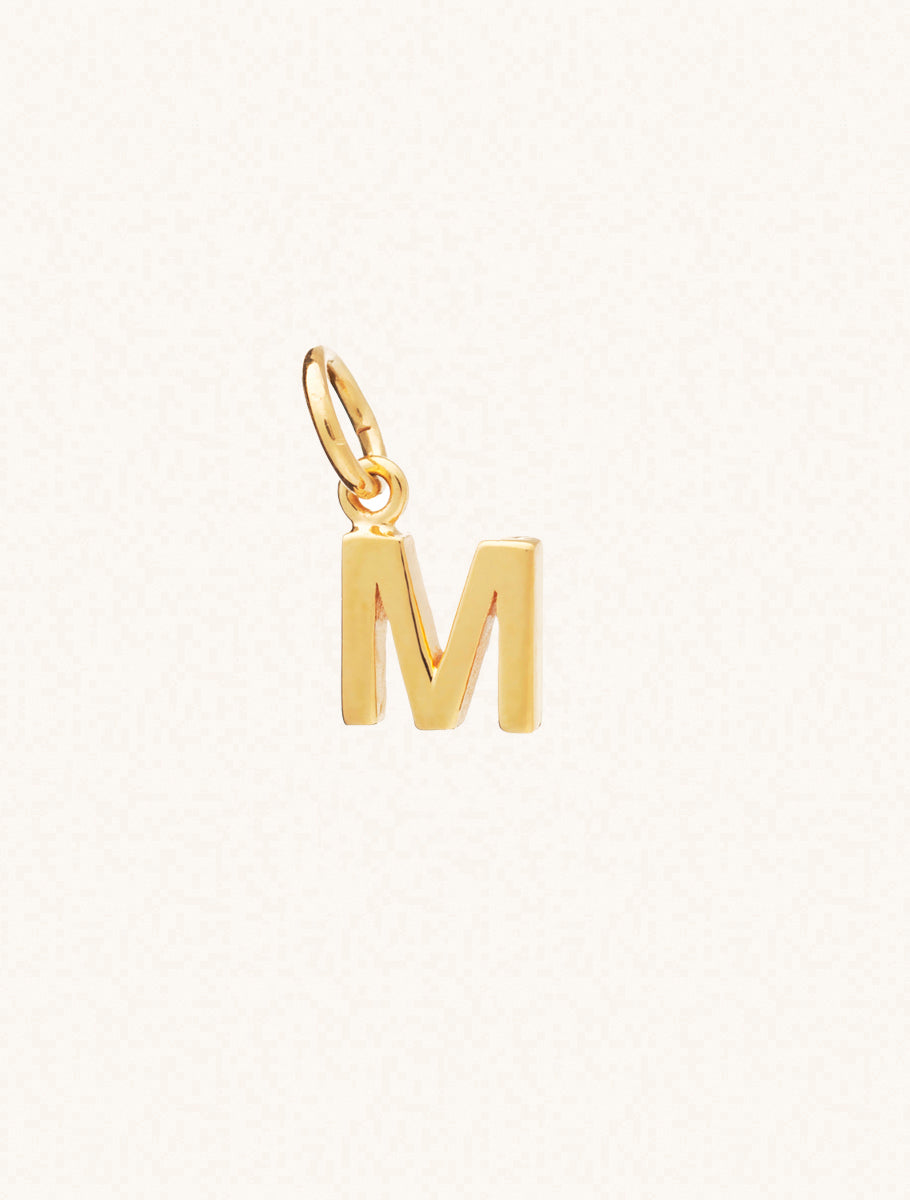 A gold caplital letter M approximately 8mm x 6mm with a connecting ring to thread through a necklace at the top. Set on a cream background.