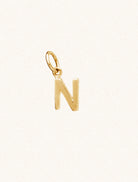 A gold caplital letter N approximately 8mm x 6mm with a connecting ring to thread through a necklace at the top. Set on a cream background.