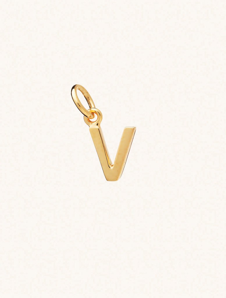 A gold caplital letter V approximately 8mm x 6mm with a connecting ring to thread through a necklace at the top. Set on a cream background.