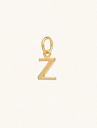 A gold caplital letter Z approximately 8mm x 6mm with a connecting ring to thread through a necklace at the top. Set on a cream background.