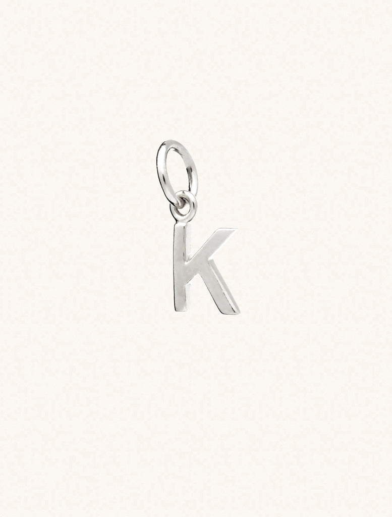 A sterling silver caplital letter K approximately 8mm x 6mm with a connecting ring to thread through a necklace at the top. Set on a cream background.